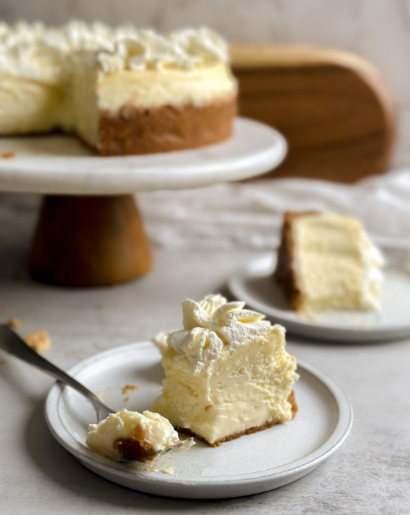 Close-up of a cheesecake slice placed sideways on a small plate, featuring a delicate crust and rich creamy texture, with another slice and a full cheesecake visible behind.