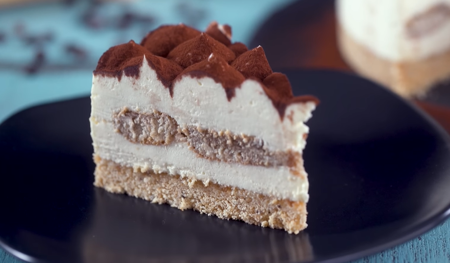 A slice of decadent baked tiramisu cheesecake rests on a small plate, featuring a rich chocolate crust, a creamy cheesecake interior, and layers of coffee-soaked ladyfingers nestled within. Atop the cheesecake are dollops of mascarpone cream, delicately dusted with cocoa powder. In the background, another slice of cheesecake adds to the tempting display, while a fork is poised to cut out a delightful bite.