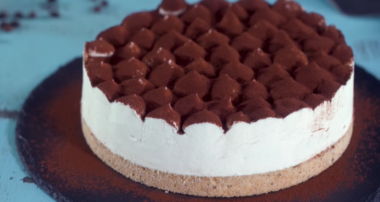 The image depicts a tiramisu cheesecake displayed on a cake stand. It showcases a chocolate crust, a plain cheesecake filling, and is topped with mascarpone cream and cocoa powder.