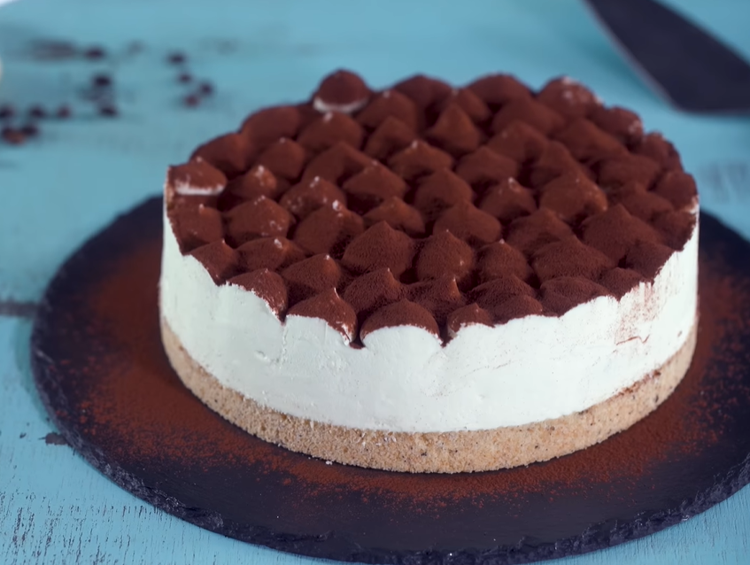 The image depicts a tiramisu cheesecake displayed on a cake stand. It showcases a chocolate crust, a plain cheesecake filling, and is topped with mascarpone cream and cocoa powder.