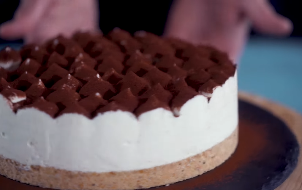 A tiramisu cheesecake presented on a cake stand, featuring a chocolate crust, a smooth cheesecake filling, and adorned with dollops of mascarpone cream lightly dusted with cocoa powder. Partial top view shown in the image.