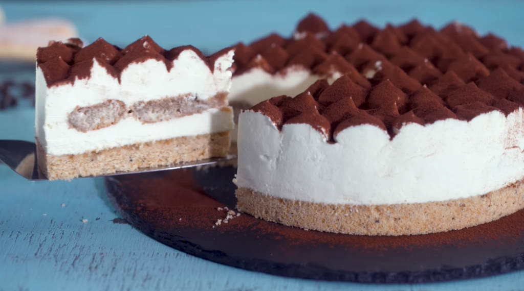 A tiramisu cheesecake displayed on a  cake stand. The cheesecake features a chocolate crust, a creamy plain cheesecake filling, and is garnished with mascarpone cream dollops dusted with cocoa powder. A spatula slices through, revealing the creamy interior and the layer of coffee-soaked ladyfingers in the middle.