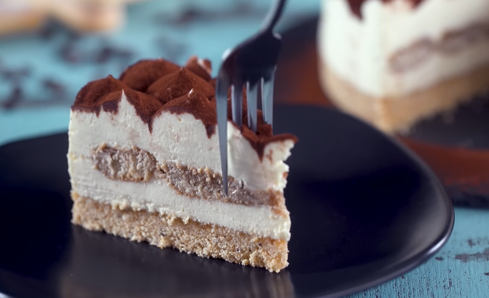 "A slice of baked tiramisu cheesecake on a plate, with chocolate crust, creamy cheesecake, coffee-soaked ladyfingers, mascarpone cream, and a fork cutting a bite."





