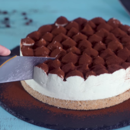 A tiramisu cheesecake is elegantly displayed on a cake stand. This dessert boasts a chocolate crust, a smooth plain cheesecake filling, and is beautifully finished with dollops of mascarpone cream dusted with cocoa powder. In a close-up image, a spatula delicately lifts a slice, revealing the creamy interior and layers of coffee-soaked ladyfingers nestled within.