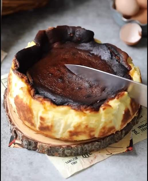 A slice of Basque cheesecake with a dark crust is being pulled out, revealing its creamy interior, all set on parchment paper.





