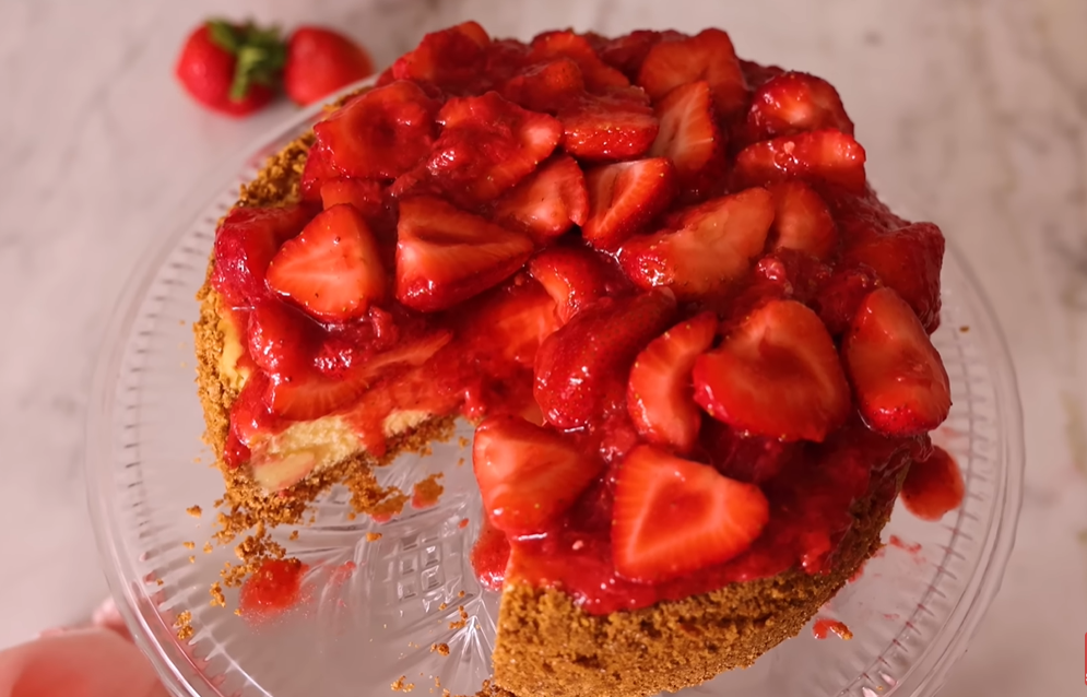 Delicious strawberry cheesecake on a serving stand topped with fresh strawberry compote and whole strawberries. A slice is being served, showcasing its creamy interior.






