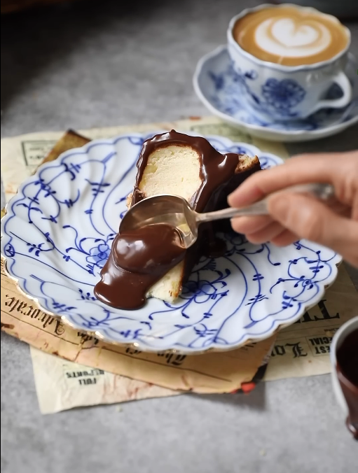 A slice of Basque cheesecake on a small plate, topped with chocolate ganache and a bite taken out with a small spoon.






