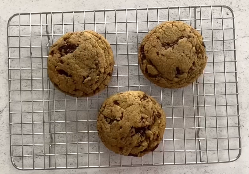 Here's an overhead picture of 6 large, thick chocolate chip cookies on a wire rack. The cookies are golden brown with chocolate chips visibly studded throughout. They are evenly spaced on the wire rack, which rests on a rustic wooden table background