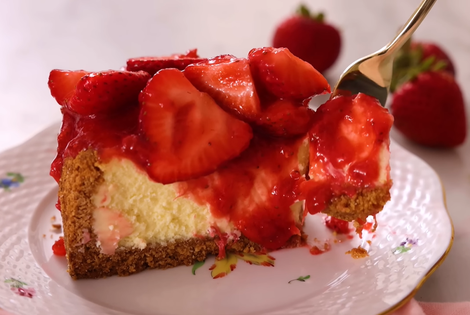 Slice of cheesecake on a small plate with a fork taking a bite, featuring a creamy texture and topped with saucy strawberry compote.



