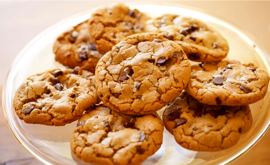 A plate of brown chocolate chip cookies with crisp edges and soft, chewy interiors. The cookies are generously studded with semi-sweet chocolate chips and have slightly cracked surfaces, making them look delicious and inviting.