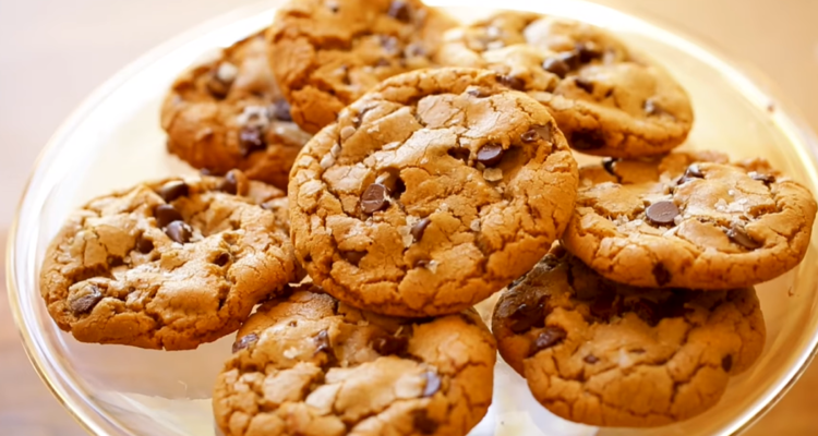 A plate of golden-brown chocolate chip cookies with crisp edges and soft, chewy interiors. The cookies are generously studded with semi-sweet chocolate chips and have slightly cracked surfaces, making them look delicious and inviting.