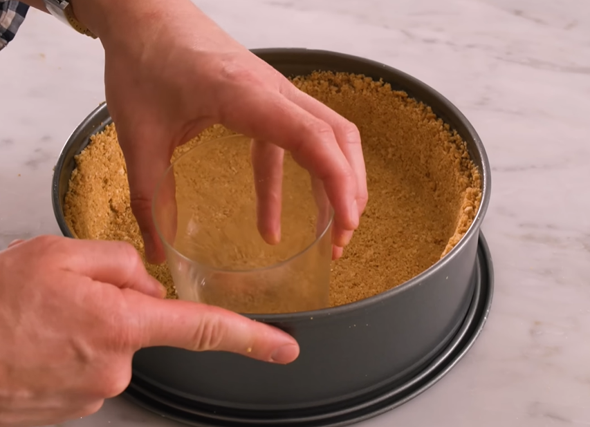 Cheesecake crust ingredients in a springform pan being pressed down by a hand using a measuring cup.






