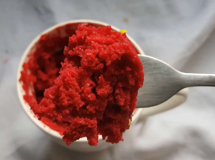 A top-down view of a  mug filled with rich, red velvet cake topped with a swirl of creamy, smooth cream cheese frosting. A spoon lifts a moist, fluffy bite, revealing the cake’s soft texture and deep red hue.