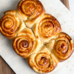 Six freshly baked cheesy bread rolls in a line on a serving board, featuring a rich golden color and a drizzle of honey butter on top.